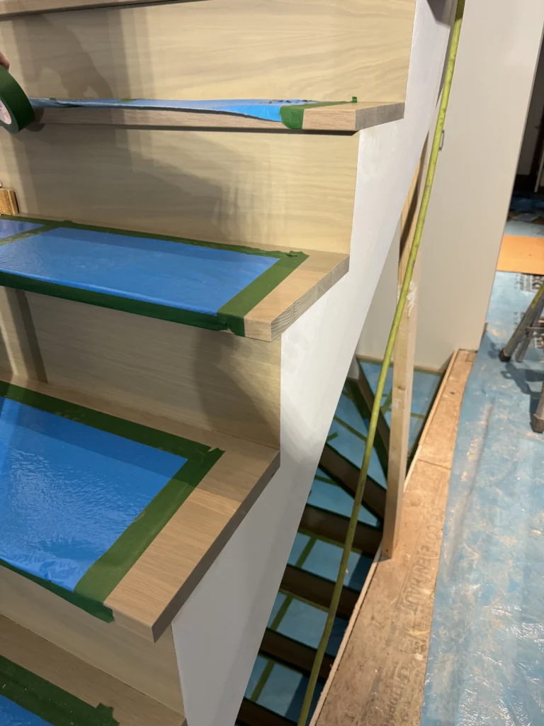 Escalier en chêne blanc en cours de finition, marches protégées par un film bleu et un ruban pendant la peinture blanche des contremarches