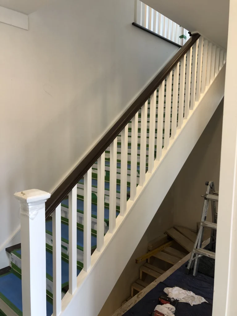 Escalier droit à barreaux blancs et main courante de bois teinté brun, en cours de finition